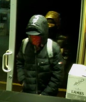 Suspect 01 entering the store. They are wearing a black coat with hood up and wearing a backpack with grey straps. 